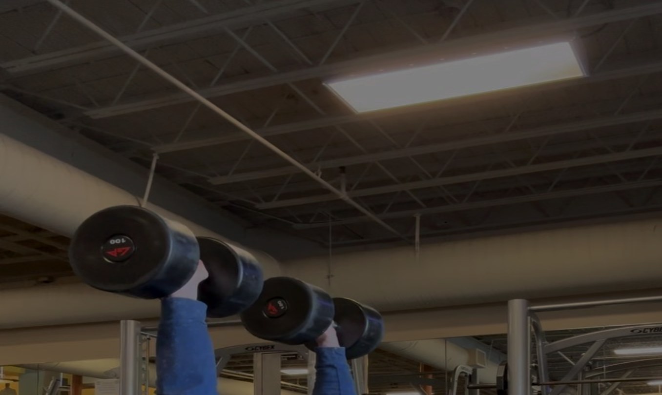 Brandon Kolar performing a dumbbell overhead press under gym lighting
